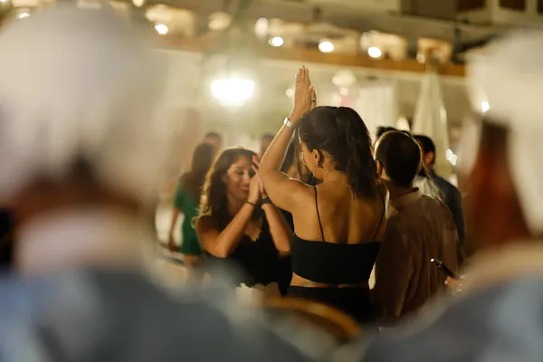 Two women are dancing together in a lit room, surrounded by other people socializing.