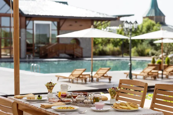 Table de petit-déjeuner en terrasse avec nourriture, boissons et fruits frais, vue sur une piscine avec des transats et parasols