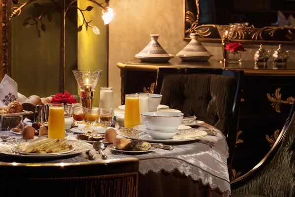 Table set for breakfast with drinks, eggs, pastries, and fruit, in an elegant setting with candelabras and cushions.
