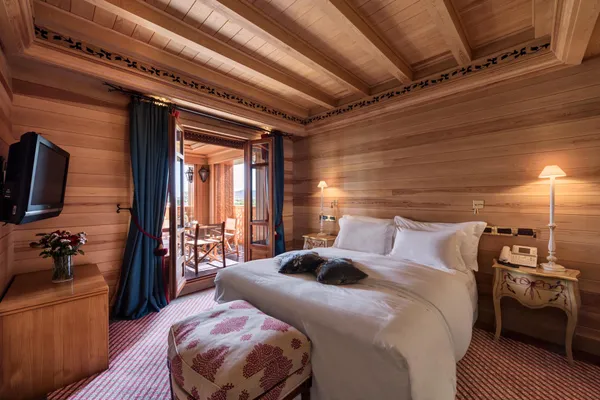 Wooden room with a double bed, a television, a bedside table with a lamp and a phone, and a French window opening onto a balcony.