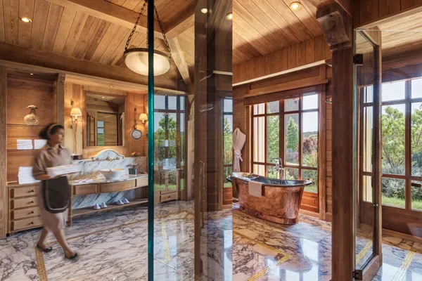 A person walks in a wooden bathroom with a copper bathtub, a marble sink, and large windows overlooking the outside.