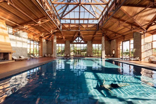 Indoor pool with a wooden roof and large windows, surrounded by lounge chairs and stone columns