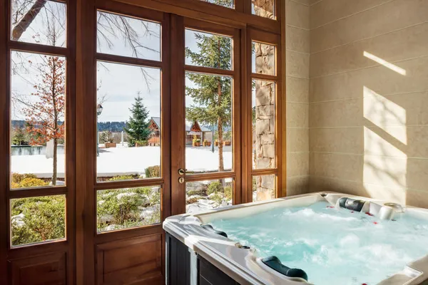 Jacuzzi filled with water with a view of a snowy landscape through large wooden windows
