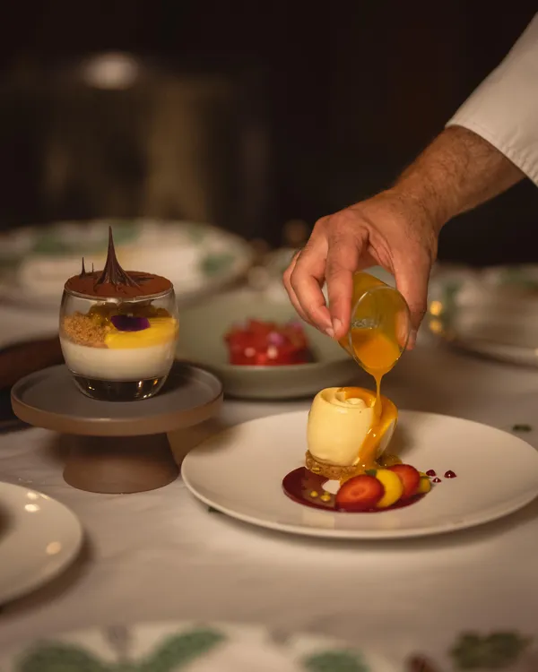 Une main verse un liquide doré sur une préparation culinaire posée sur une assiette blanche