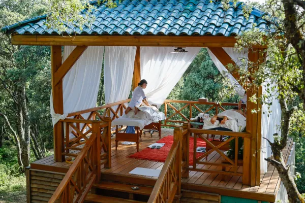 Two people lying on outdoor massage tables under a wooden awning with a blue-tiled roof, surrounded by trees.