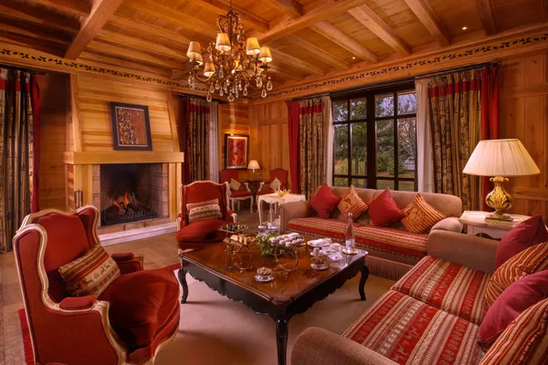 Wooden living room with lit fireplace, red sofas and armchairs, coffee table with tea and candelabras, chandelier and large windows