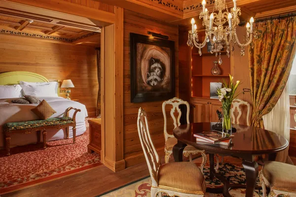 Wooden room with bed, chandelier, painting, table with chairs, and flowers in a vase.